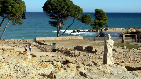 Pla general del jaciment d'Empúries, amb el mar al fons, i dos visitants passejant per les ruïnes Pla general del jaciment d'Empúries, amb el mar al fons, i dos visitants passejant per les ruïnes