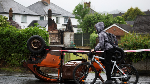Una persona pasa en bicicleta junto a un coche quemado frente a una casa abandonada e incendiada en Antrim Road en los disturbios de Belfast. Una persona pasa en bicicleta junto a un coche quemado frente a una casa abandonada e incendiada en Antrim Road en los disturbios de Belfast.