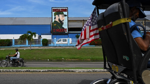 Un bicitaxi decorado con la bandera de Estados Unidos y un hombre en motocicleta pasan junto a un cartel del difunto presidente cubano Fidel Castro.