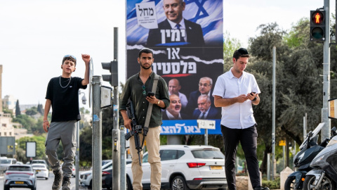 Un hombre israelí armado espera junto a otras personas para cruzar un cruce cerca de una valla publicitaria de la campaña electoral del partido Likud de Benjamin Netanyahu, en Jerusalén, en octubre de este año