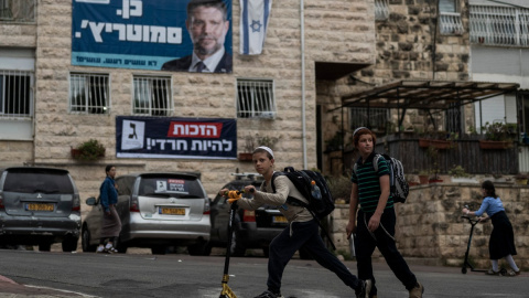 Unos niños pasan junto a un edificio residencial adornado con pancartas electorales de los partidos Sionismo Religioso (arriba) y Judaísmo Unido de la Torá (abajo) en Jerusalén, en la víspera de las elecciones al Knéset