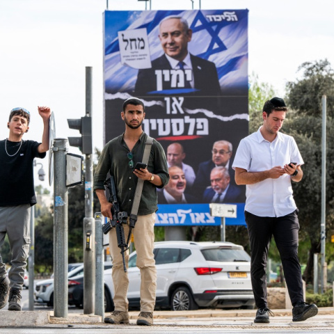 Un hombre israelí armado espera junto a otras personas para cruzar un cruce cerca de una valla publicitaria de la campaña electoral del partido Likud de Benjamin Netanyahu, en Jerusalén, en octubre de este año