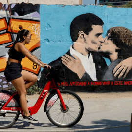 Fotografía de septiembre de 2023 de una joven en bicicleta que pasa junto a un graffiti en Barcelona que presenta a Pedro Sanchez y a Carles Puigdemont besándose. REUTERS/Albert Gea