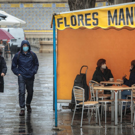 Dos personas en la terraza de un bar protegidas de la lluvia por un toldo de un puesto de flores.