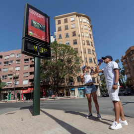 Dos jóvenes beben agua junto a un termómetro que marca 43 grados centígrados, en la Plaza del Royo de Murcia.