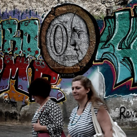 Dos mujeres caminan frente a un muro con un grafitti en el que aparece una moneda de euro con un 0 en su interior. AFP