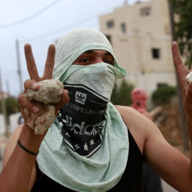 Un palestino con el rostro tapo, hace el gesto de la victoria con sus manos en las que lleva piedras, en la ciudad de Dura, cerca de Hebrón. /EFE-ABED AL HASHLAMOUN