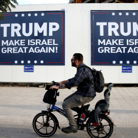 Un hombre montado en bicicleta pasa junto a unos letreros en Tel Aviv parafraseando el lema de campala de Donadl Trump. REUTERS/Baz Ratner