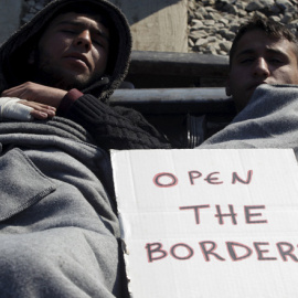 Dos refugiados descansan en el suelo cerca del pueblo de Idomeni, Grecia, con un cartel que pone "abre las fronteras"./REUTERS