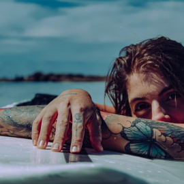 Una mujer tatuada descansa sobre una tabla de surf en la playa. Foto: Pexels