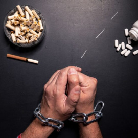 Unas manos con esposas junto a un cenicero y un bote de pastillas.