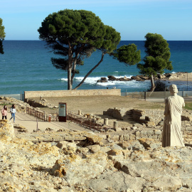 Pla general del jaciment d'Empúries, amb el mar al fons, i dos visitants passejant per les ruïnes
