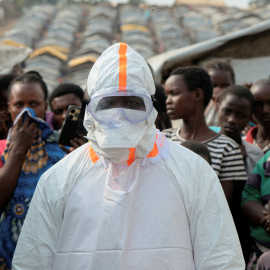 Un trabajador sanitario, equipado con equipo de protección individual (EPI), junto a un grupo de desplazados que esperan el entierro de presuntas víctimas del ébola en el campamento de Kigonze.