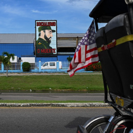 Un bicitaxi decorado con la bandera de Estados Unidos y un hombre en motocicleta pasan junto a un cartel del difunto presidente cubano Fidel Castro.