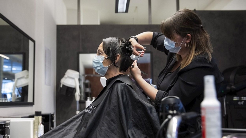 27/04/2020.- Una peluquera corta el pelo a una mujer con una máscara facial protectora contra el coronavirus en una peluquería de Berna, Suiza este lunes. Servicios como la peluquería, los masajes, los tatuajes o los salones de belleza están abiertos al p