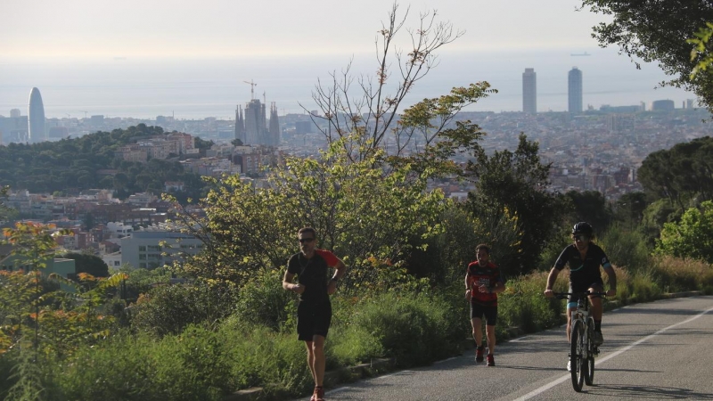 Pla general d'un ciclista i dos corredors pujant pel carrer Manuel Arnús, just abans d'arribar a la carretera de les Aigües, al Parc de Collserola de Barcelona. Imatge del 2 de maig de 2020. (Horitzontal)