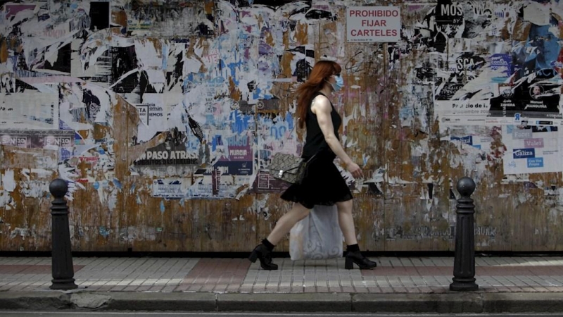 Una mujer con mascarilla camina este jueves por una céntrica calle de A Coruña, frente a un muro lleno de carteles con actividades culturales,en la jornada en la que ha entrado en vigor llevar este tipo de protección en espacios públicos, tanto al aire l