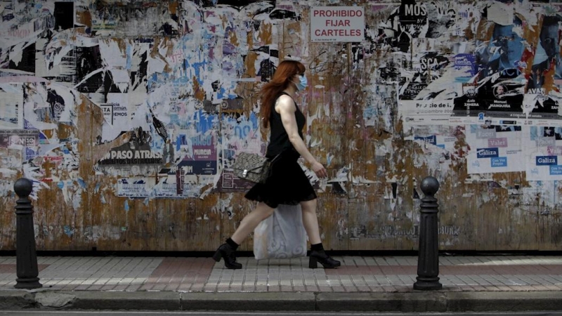 Una mujer con mascarilla camina este jueves por una céntrica calle de A Coruña, frente a un muro lleno de carteles con actividades culturales,en la jornada en la que ha entrado en vigor llevar este tipo de protección en espacios públicos, tanto al aire l