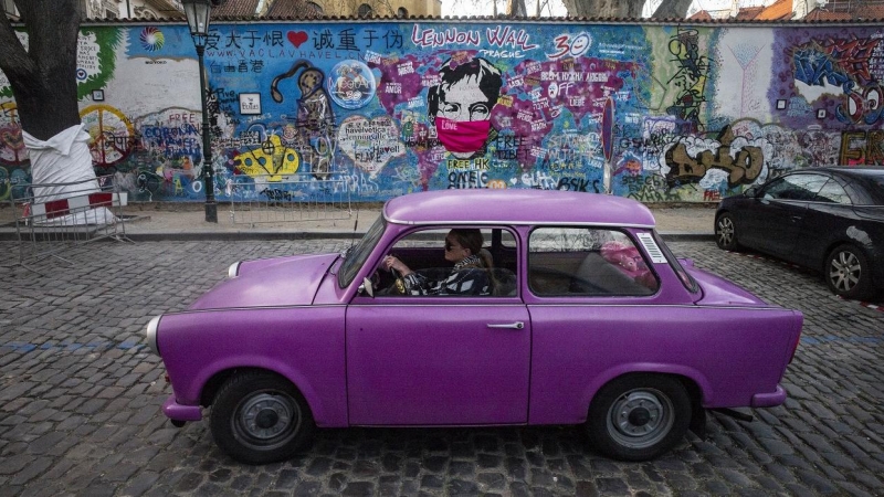 Una mujer con mascarilla conduce un coche violeta junto al llamado Muro de John Lennin, en el centro histórico de Praga. AFP/Michal Cizek