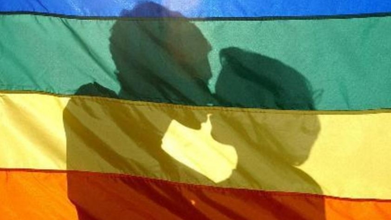 Una pareja se besa tras la bandera del arcoíris, símbolo del movimiento LGTBI.