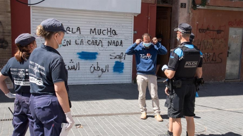 Agentes cívicos (i) y de la Guardia Urbana (d) entregan una mascarilla a un hombre que no llevaba, este jueves, cuando se han puesto en marcha las medidas especiales de restricción de movimientos. /EFE