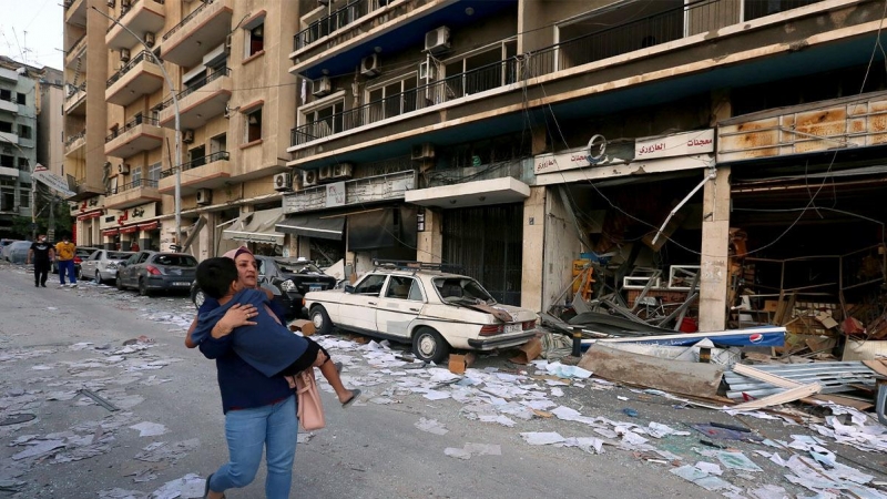 Una mujer lleva a un niño mientras pasa por las tiendas dañadas tras la explosión del martes en Beirut, Líbano