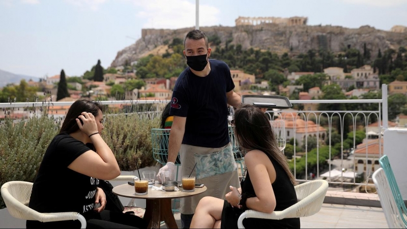 ​Un camarero con mascarilla atiende a unas clientas en una terraza de Atenas, con la Acrópolis al fondo. REUTERS/Costas Baltas