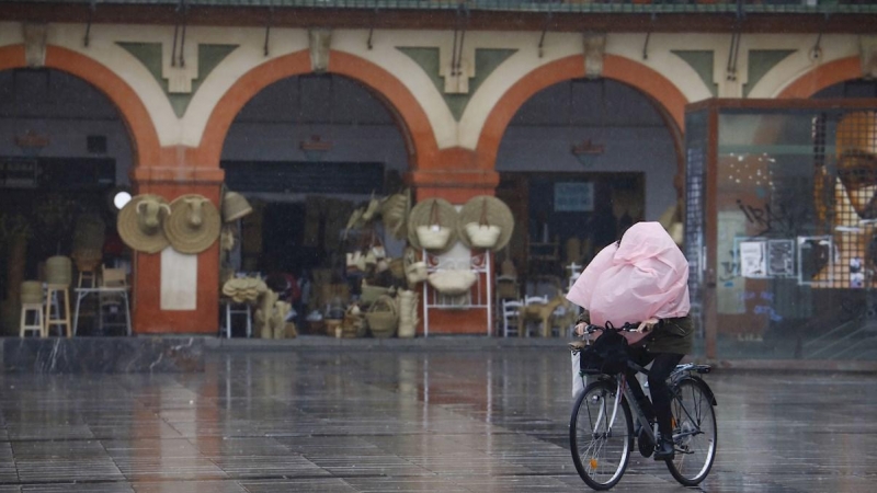 Una mujer en bicicleta a la que el viento le ha tapado la cara totalmente con el chubasquero mientras pasa por la Plaza de la Corredera de Córdoba