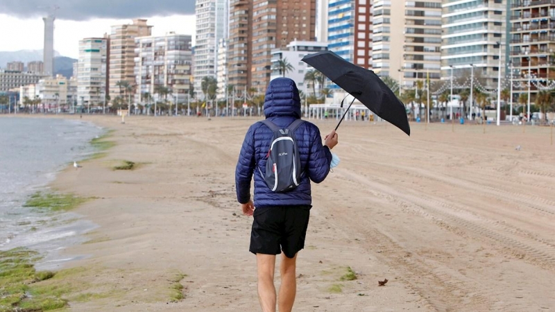 Una persona se protege de la lluvia con un paraguas mientras pasea por la playa de Levante de Benidorm