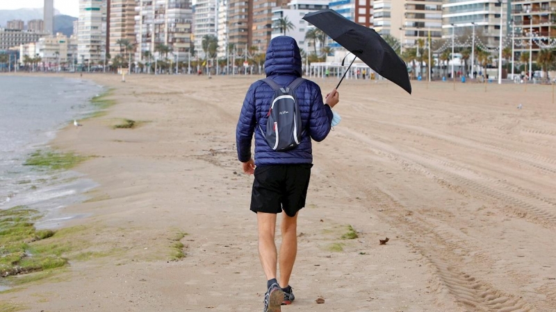 Una persona se protege de la lluvia con un paraguas mientras pasea por la playa de Levante de Benidorm