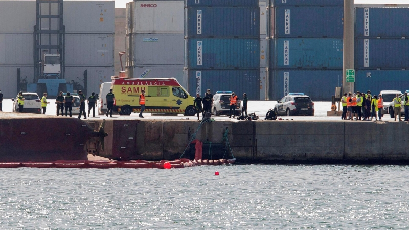 Efectivos de la Guardia Civil y del servicio de emergencias esperan en el Muelle del Centenario de Port Castelló durante las tareas de búsqueda de dos personas desaparecidas tras el vuelco del buque 'Nazmuye Ana' (a la izquierda, en la imagen), este sábad