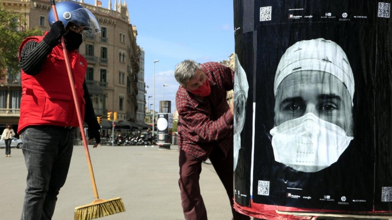 Cartells amb cares de professionals sanitaris amb mascareta en una columna publicitària de plaça de Catalunya de Barcelona, en el projecte 'Behind the mask', que busca el reconeixement col·lectiu davant de la covid-19. 8 d'abril del 2021.