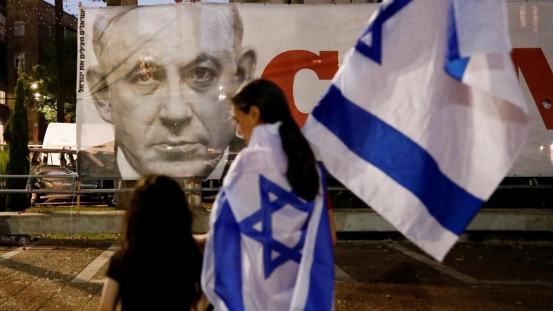 Personas con banderas de Israel pasan por delante de un cartel con la imagen de Benjamin Netanyahu en Tel Aviv, después de la votación en el Parlamento a un nuevo Gobierno de coalición, que pone fin a sus 12 años en el poder. REUTERS/Corinna Kern