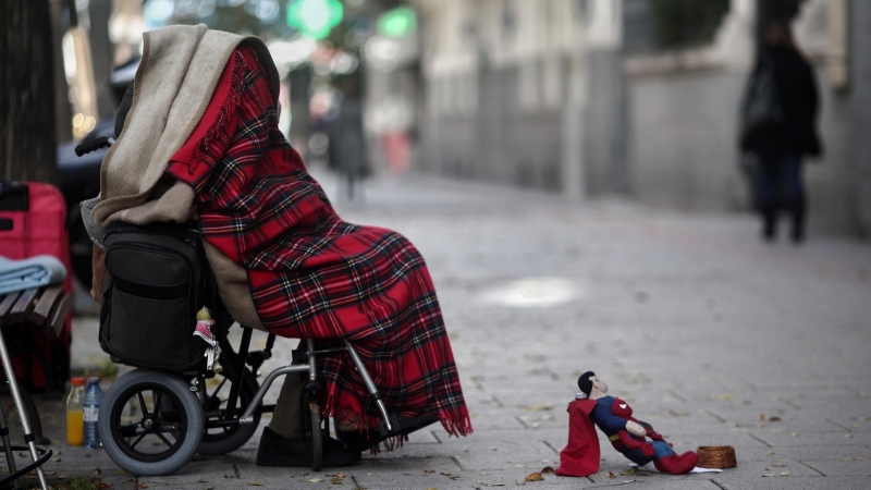 Una persona en silla de ruedas y tapada con una manta pide dinero con un muñeco de Superman en una céntrica calle.