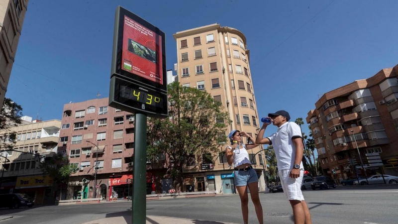 Dos jóvenes beben agua junto a un termómetro que marca 43 grados centígrados, en la Plaza del Royo de Murcia.