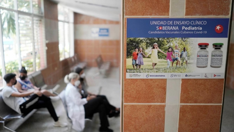 Imagen de archivo de varias personas sentadas cerca de un cartel que anuncia los ensayos clínicos de la vacuna Soberana 02, en una clínica sanitaria en La Habana. - REUTERS