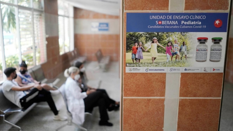 Imagen de archivo de varias personas sentadas cerca de un cartel que anuncia los ensayos clínicos de la vacuna Soberana 02, en una clínica sanitaria en La Habana. - REUTERS