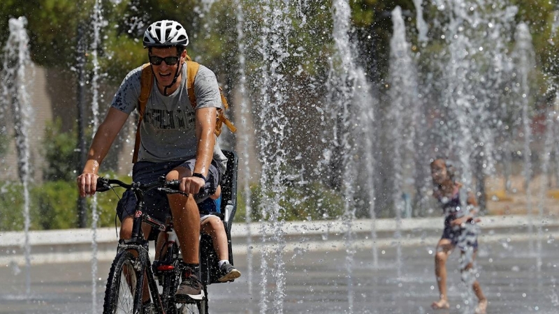 Un padre con su hijo en bicicleta se refrescan en las fuentes del Parque Central de València. - EFE