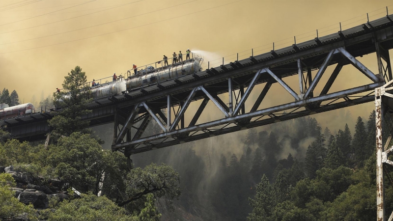 Los bomberos trabajan apagando el fuego sobre un tren con un tanque de agua , en ese momento atraviesan un puente.