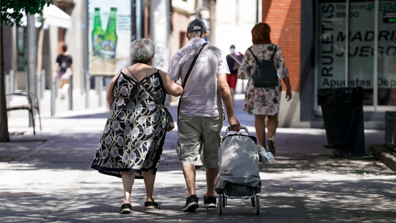 Una pareja de ancianos camina por la calle con un carro de compra.