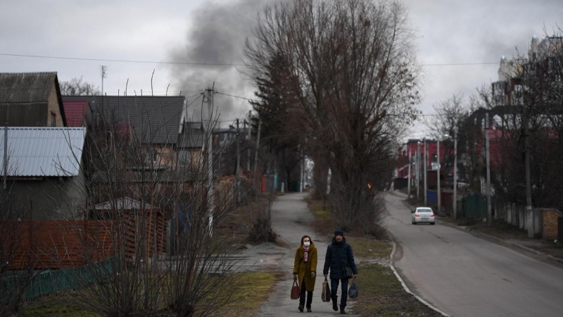 Población local camina por una calle mientras el humo se eleva cerca de la ciudad de Hostomel y el aeropuerto Antonov, en el noroeste de Kiev, donde fuerzas rusas y ucranianas luchan por el control de una base aérea en las afueras del norte de la capital