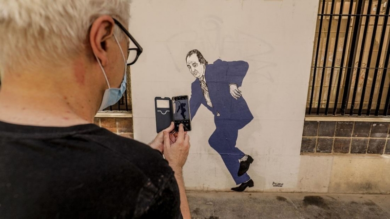 Un hombre hace una fotografía con su móvil al graffiti del rey emérito Juan Carlos I tropezando y a punto de caer, obra pintada en una céntrica calle de Valencia por el artista gallego apodado 'el Primo de Banksy', en Valencia, Comunidad Valenciana (Españ