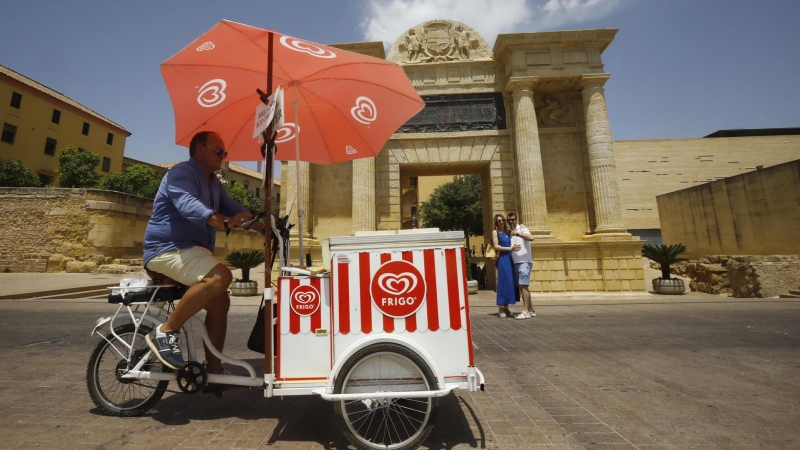 Un vendedor de helados protegido con una sombrilla, pasa con su bicicleta junto la puerta del puente de Córdoba