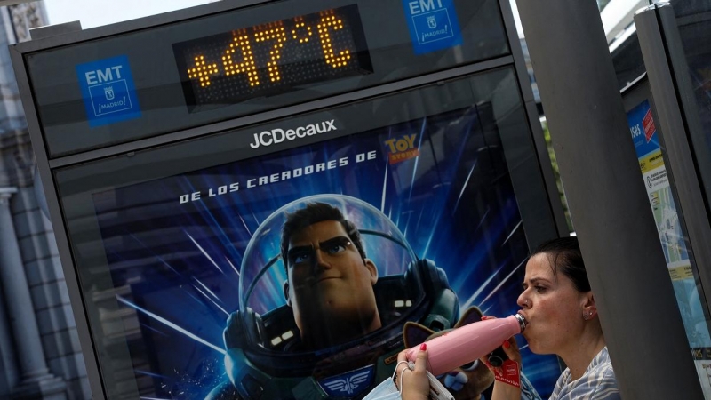 Una mujer bebe agua en la sombra de una parada de autobús de Madrid cuyo termómetro marca 47ºC.