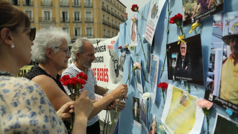 (22/5/22) Varias personas colocan claveles en un mural con retratos de colaboradores en la búsqueda de víctimas por el robo de bebés en España, a 22 de mayo de 2022.