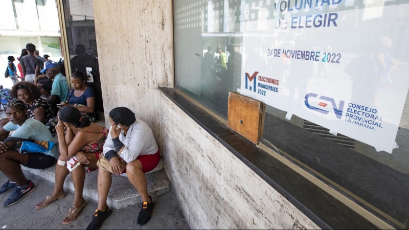 Varias personas permanecen sentadas junto a un cartel de las elecciones locales del 27 de noviembre.