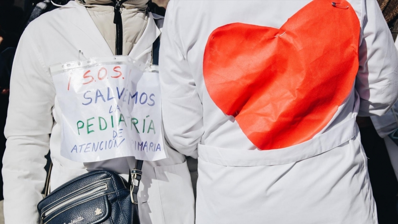 Dos personas protestan con un cartel que reza '¡S.O.S.! Salvemos la pediatría de Atención Primaria' y con un dibujo de un corazón pegado a la espalda durante una concentración de médicos y pediatras de Atención Primaria en huelga, entre la sede de la Dire