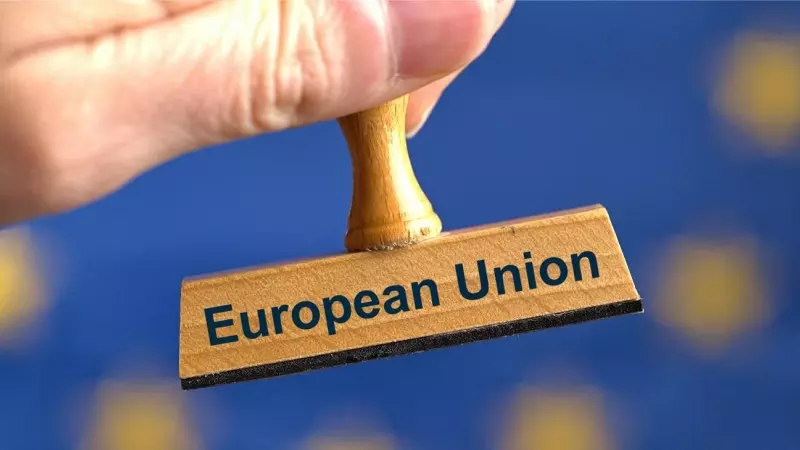Un sello de madera simbólico con la inscripción 'Unión Europea', sostenido por una mano frente a una bandera borrosa de la UE en el fondo.