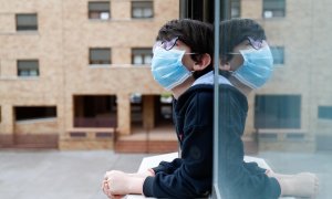 Un niño con una mascarilla se asoma a la ventana de su casa. EP