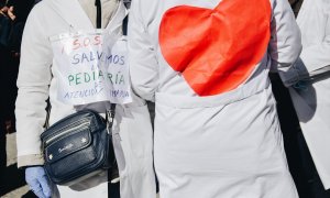 Dos personas protestan con un cartel que reza '¡S.O.S.! Salvemos la pediatría de Atención Primaria' y con un dibujo de un corazón pegado a la espalda durante una concentración de médicos y pediatras de Atención Primaria en huelga, entre la sede de la Dire